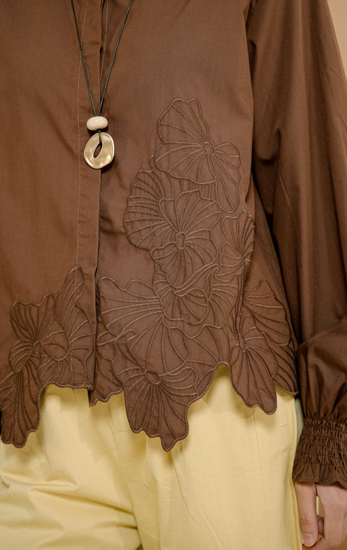 Model mengenakan Iridessa Embroidered Button-Up Shirt Dark Brown, kemeja katun wanita lengan panjang warna cokelat tua dengan detail bordir bunga dan ujung keliman bergelombang. 