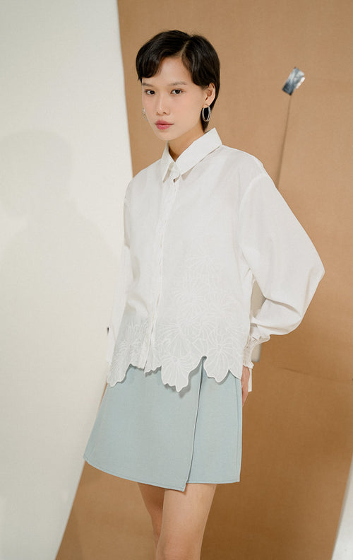 Model berdiri mengenakan Iridessa Embroidered Button-Up Shirt White, kemeja katun wanita lengan panjang warna putih dengan detail bordir bunga dan ujung keliman bergelombang. 