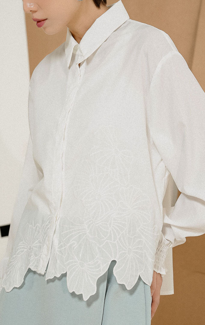 Model berdiri mengenakan Iridessa Embroidered Button-Up Shirt White, kemeja katun wanita lengan panjang warna putih dengan detail bordir bunga dan ujung keliman bergelombang. 