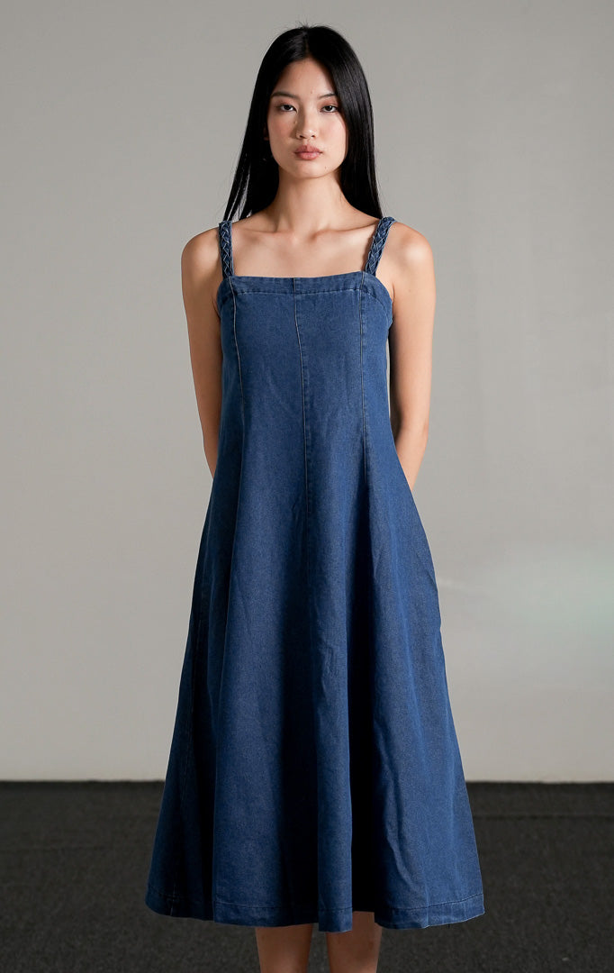 Iriska Dress berwarna indigo (biru gelap), sebuah midi dress berbahan denim tanpa lengan dengan kerah kotak, siluet A-line, dan tali bahu bertekstur, dipadukan dengan sandal hitam.