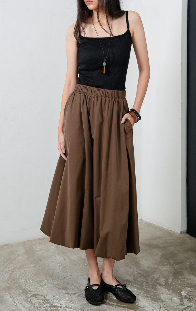 Rok panjang wanita mengalir (flowy long skirt) warna cokelat (brown) Irwin Skirt dengan siluet balon halus (subtle balloon silhouette), pinggang karet elastis yang nyaman (comfortable stretch waistband), dan saku samping (side pockets).