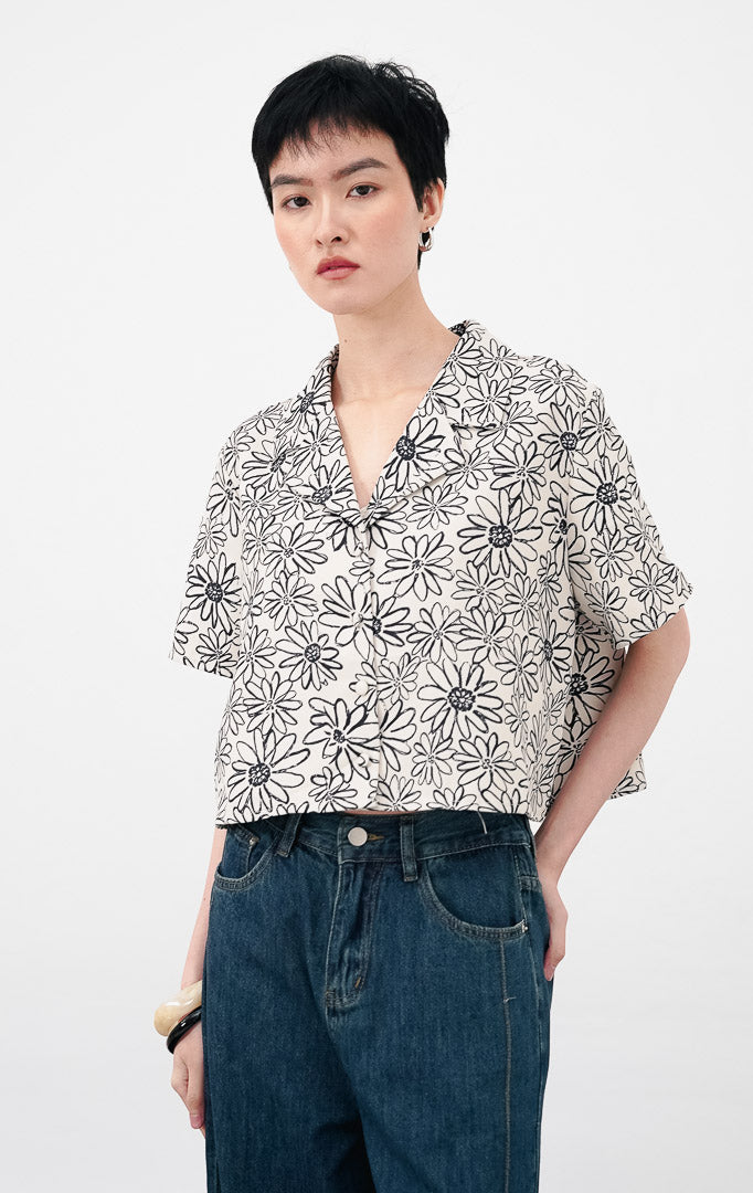Model mengenakan Isadora Top warna ivory, atasan crop lengan pendek berkerah dengan motif bunga (floral) hitam dan kancing depan, dipadukan dengan celana denim.