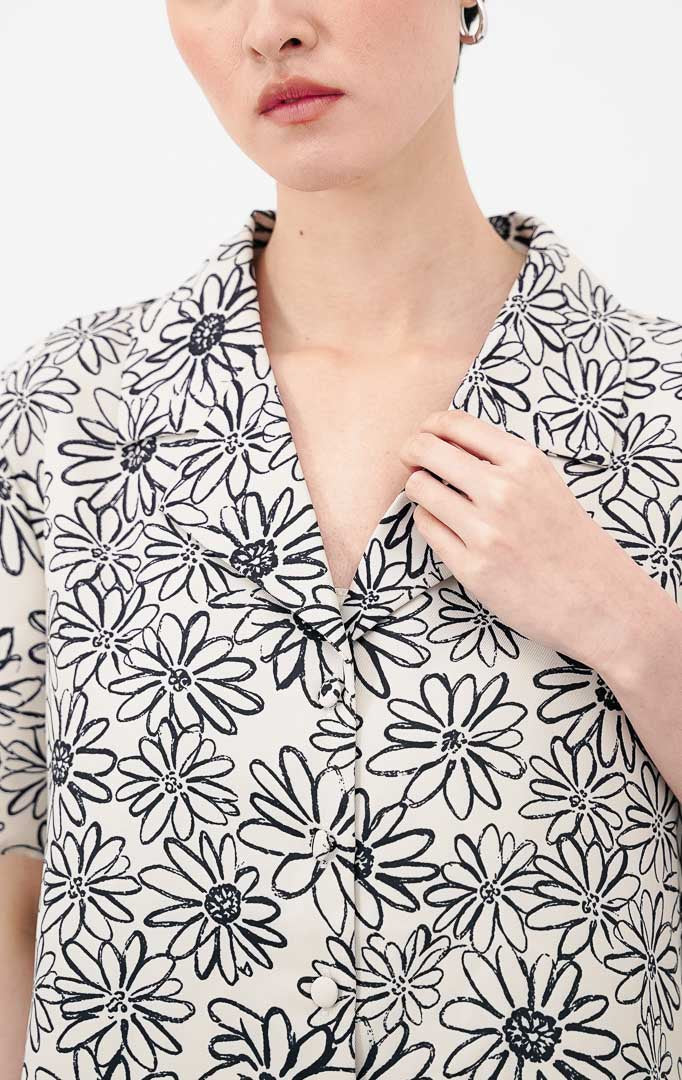 Model mengenakan Isadora Top warna ivory, atasan crop lengan pendek berkerah dengan motif bunga (floral) hitam dan kancing depan, dipadukan dengan celana denim.