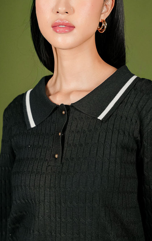 Sweater rajut wanita Isyana Sweater warna hitam lengan panjang, dilengkapi kerah polo beraksen garis putih, 3 kancing depan berwarna emas, tekstur rajut kepang (cable-knit) vertikal, dan aksen dua garis putih di ujung bawah.