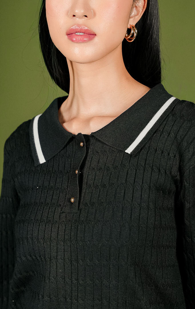 Sweater rajut wanita Isyana Sweater warna hitam lengan panjang, dilengkapi kerah polo beraksen garis putih, 3 kancing depan berwarna emas, tekstur rajut kepang (cable-knit) vertikal, dan aksen dua garis putih di ujung bawah.