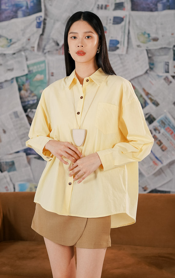 Model wanita mengenakan Izumi Top Yellow, sebuah kemeja lengan panjang model oversized warna kuning pastel (kuning muda) dengan detail kancing depan cokelat dan satu saku dada.1
