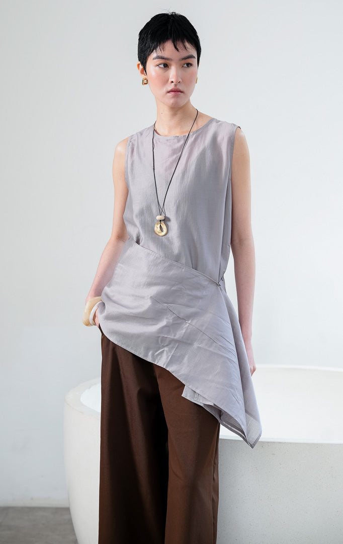 Atasan wanita tanpa lengan bahan kain halus ringan warna abu-abu (grey sleeveless top) Jaide Top dengan kerah bulat sederhana dan detail lapisan luar lilitan yang dapat dilepas pasang di bagian pinggang (detachable wrap layer at the waist). 