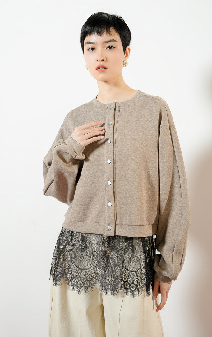 Model wanita mengenakan Oulia Jacket warna taupe, jaket cardigan rajut jersey wanita berwarna cokelat dengan kancing jepret metalik dan detail kelim renda brokat hitam transparan yang anggun. 
