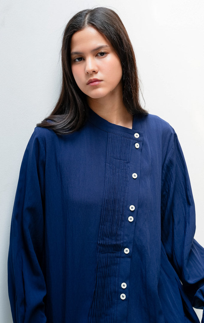 Blus wanita lengan panjang warna biru dongker (navy) Janessa Top dengan kerah asimetris (asymmetrical neckline), detail lipit pintuck vertikal di bagian depan, dan bukaan kancing tersembunyi (concealed button placket).