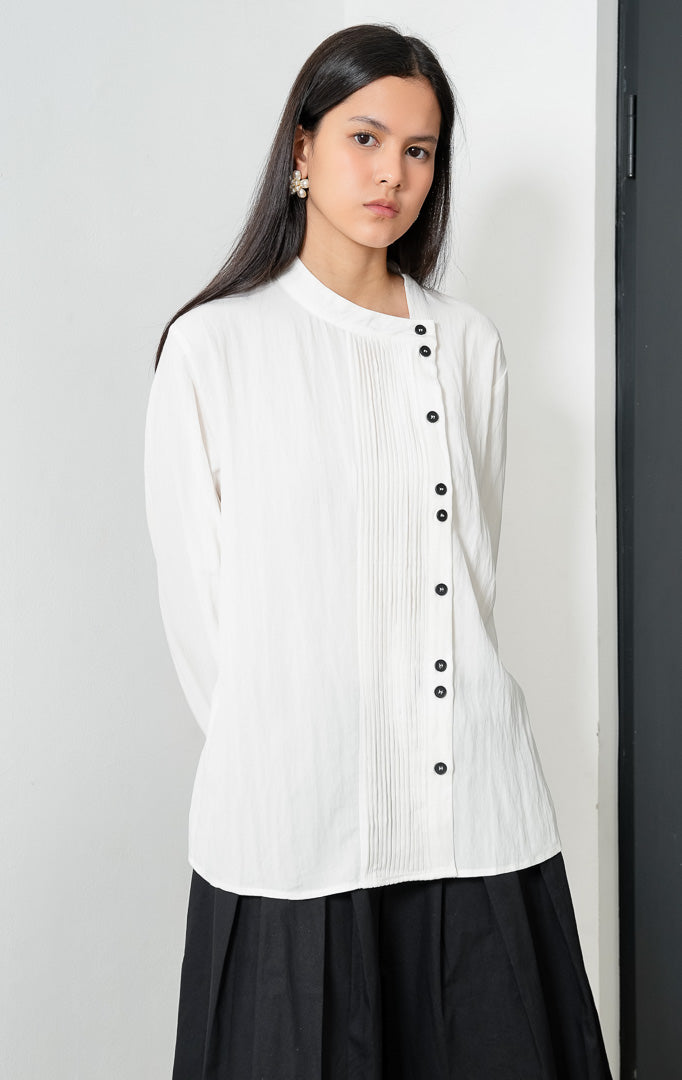 Blus wanita lengan panjang warna putih (white) Janessa Top dengan kerah asimetris (asymmetrical neckline), detail lipit pintuck vertikal di bagian depan, dan bukaan kancing tersembunyi (concealed button placket). 