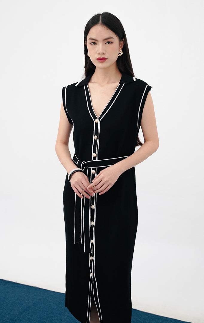 Model mengenakan Janice Dress warna black, gaun rajut tanpa lengan dengan kerah V, detail kancing emas di bagian depan, aksen garis putih vertikal, dan ikat pinggang senada.