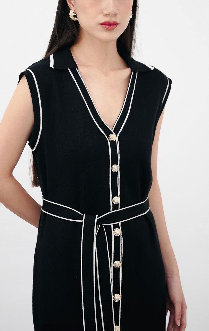 Model mengenakan Janice Dress warna black, gaun rajut tanpa lengan dengan kerah V, detail kancing emas di bagian depan, aksen garis putih vertikal, dan ikat pinggang senada.