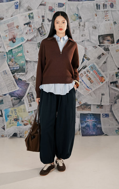 Wanita mengenakan Jett Sweater warna coklat polos (brown), sweater rajut lengan panjang dengan kerah resleting (half zip) dan potongan relaxed fit yang minimalis.