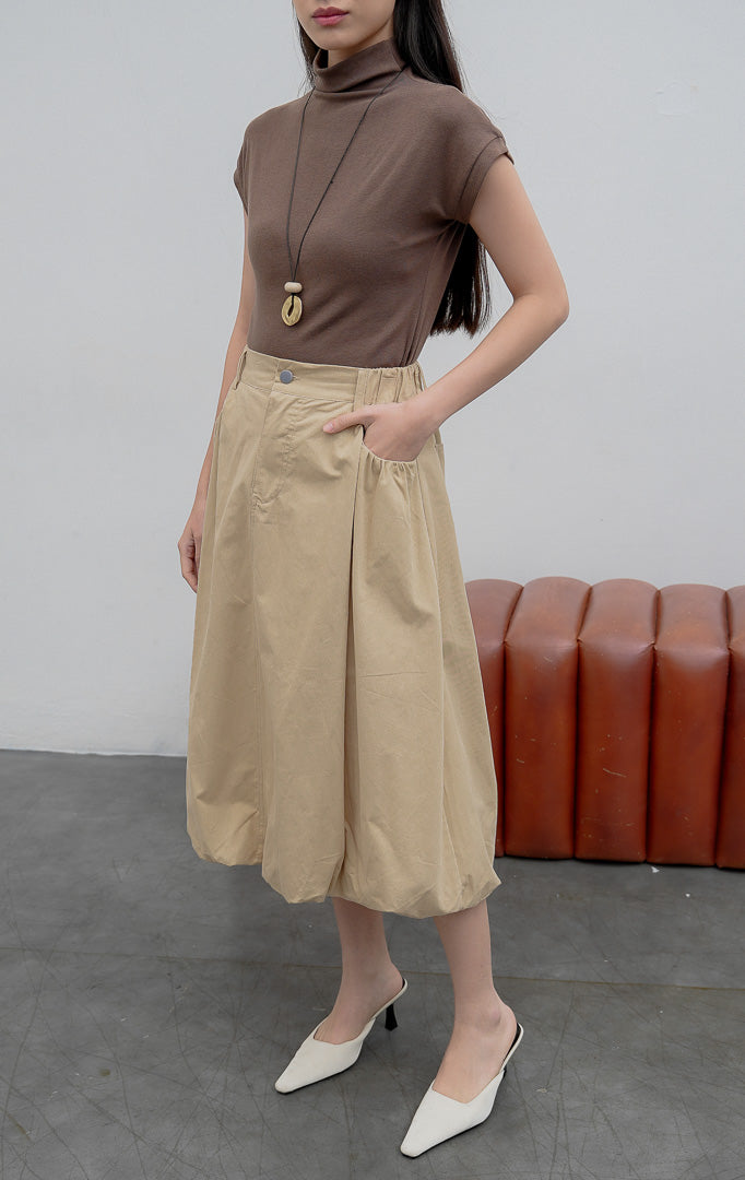 Rok balon (bubble skirt) wanita warna khaki Jinnie Skirt berbahan campuran katun-nilon terstruktur dengan siluet mekar (voluminous silhouette), detail lipitan (tucked detailing), kancing depan, dan saku samping.