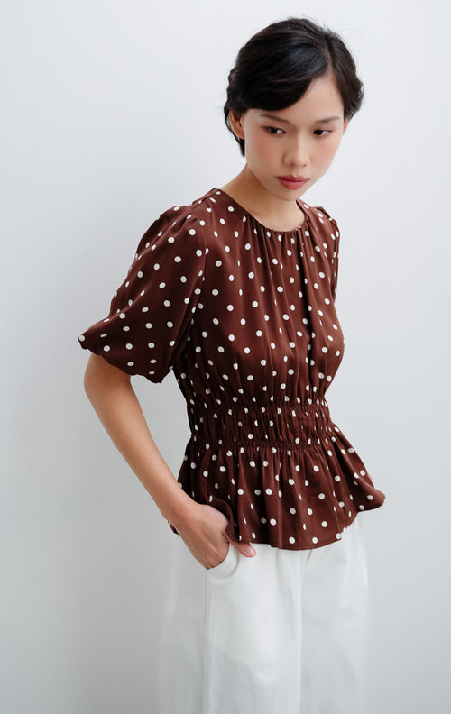 Atasan wanita warna cokelat tua (dark brown) Julietta Top bermotif polkadot dengan detail lengan balon (puff sleeves) dan badan berkerut (smocked bodice).
