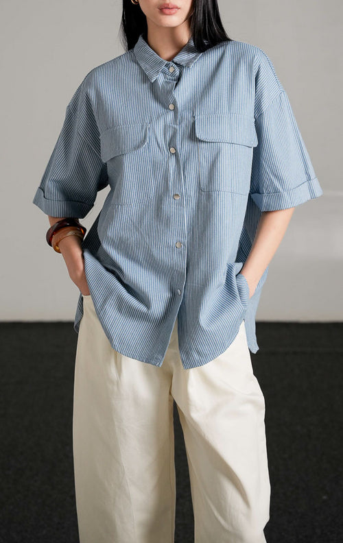 Kaine Top, kemeja denim biru motif garis tipis lengan pendek dengan saku ganda di dada, dipadukan dengan celana putih longgar.