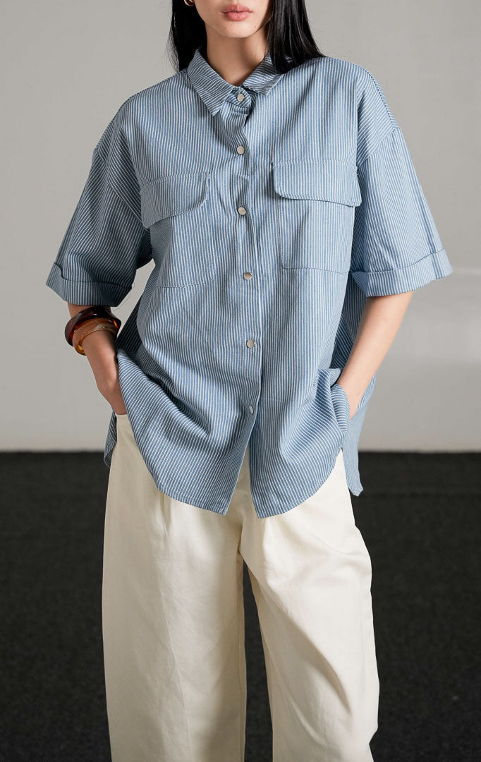 Kaine Top, kemeja denim biru motif garis tipis lengan pendek dengan saku ganda di dada, dipadukan dengan celana putih longgar.