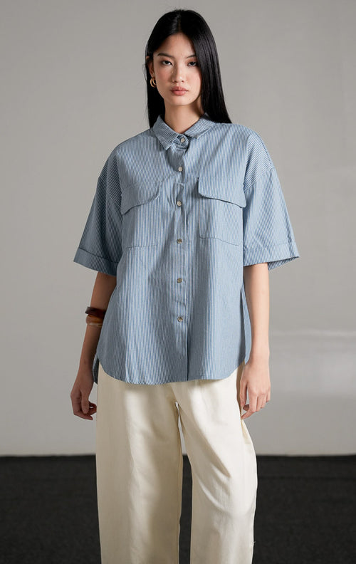 Kaine Top, kemeja denim biru motif garis tipis lengan pendek dengan saku ganda di dada, dipadukan dengan celana putih longgar.