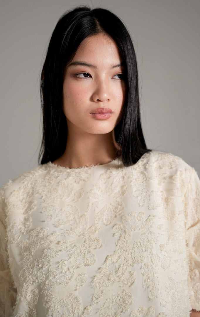 Kallani Top Ivory, atasan blus warna krem dengan tekstur fringe bulu halus dan lengan pendek semi-transparan, dipadukan dengan celana cokelat.