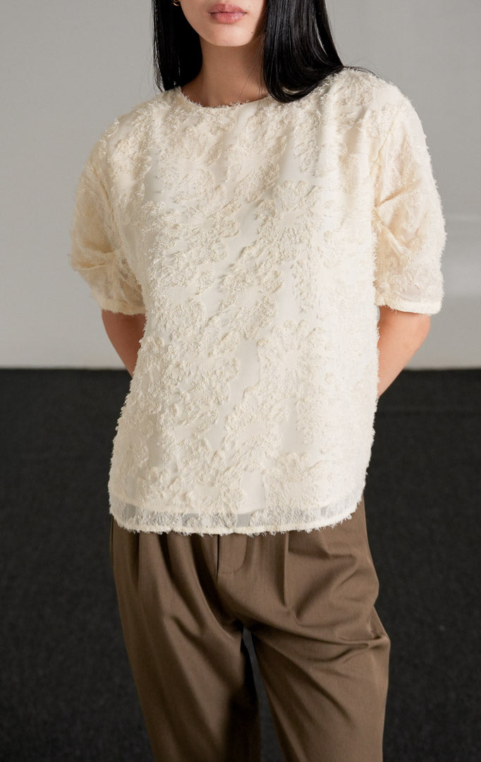 Kallani Top Ivory, atasan blus warna krem dengan tekstur fringe bulu halus dan lengan pendek semi-transparan, dipadukan dengan celana cokelat.