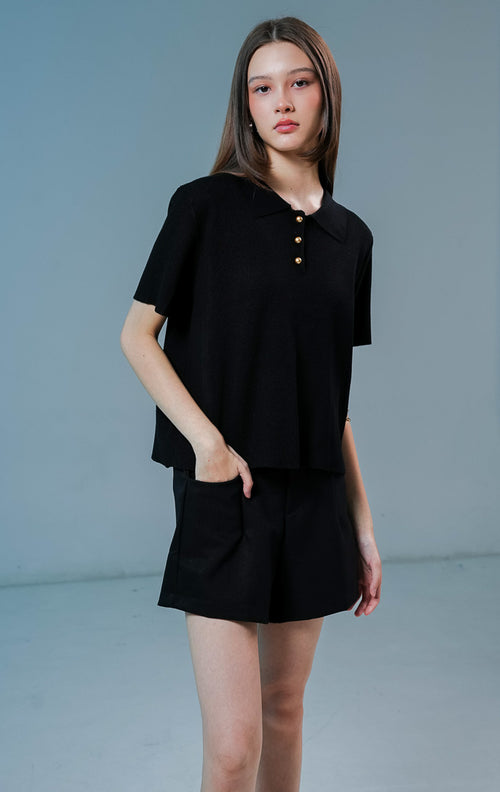 Model wanita mengenakan Griffin Collared Short Sleeve Top warna hitam, baju kaos rajut wanita lengan pendek dengan kerah terstruktur dan detail tiga kancing emas depan. 