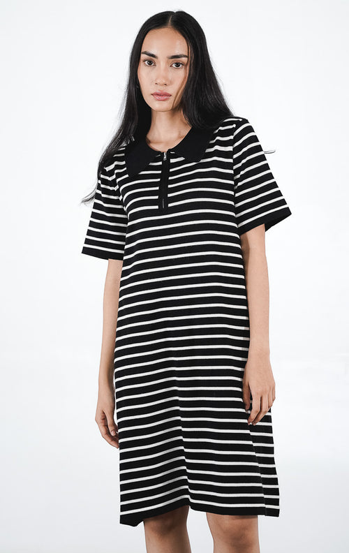 Model mengenakan Kayona Dress warna black, mini dress rajut lengan pendek motif garis horizontal putih dengan kerah polo hitam dan detail resleting depan.