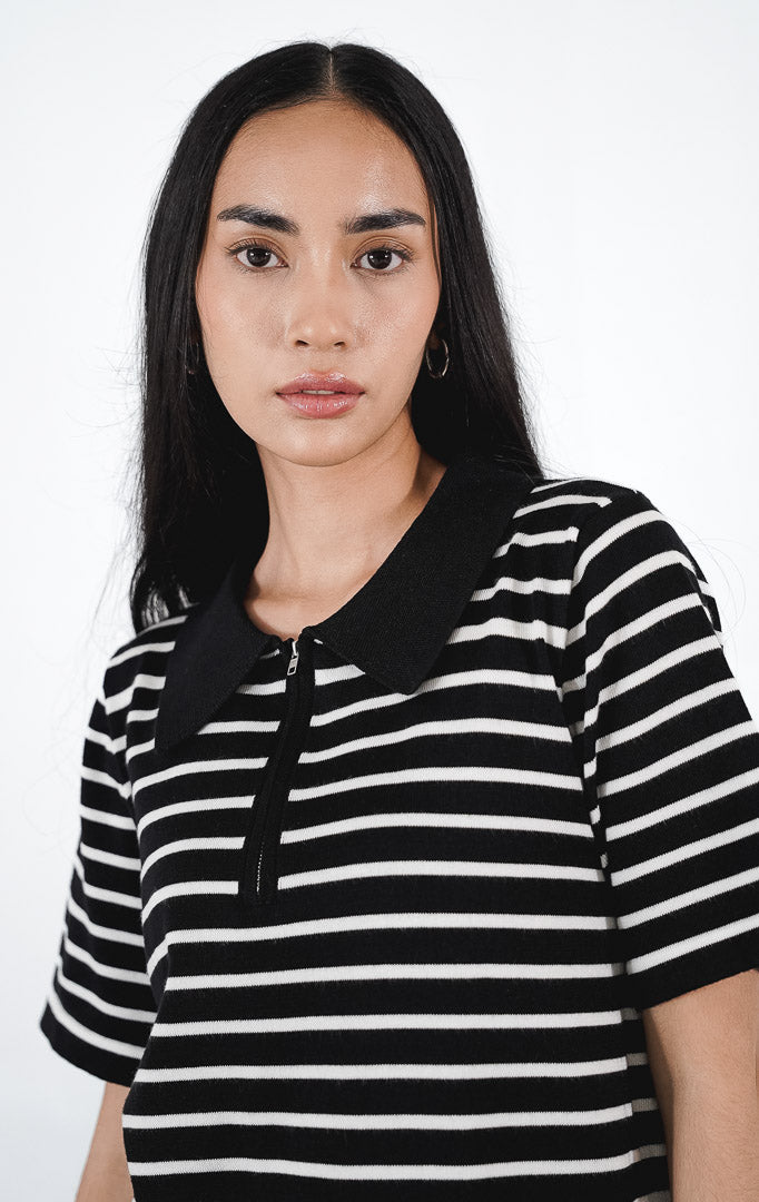 Model mengenakan Kayona Dress warna black, mini dress rajut lengan pendek motif garis horizontal putih dengan kerah polo hitam dan detail resleting depan.