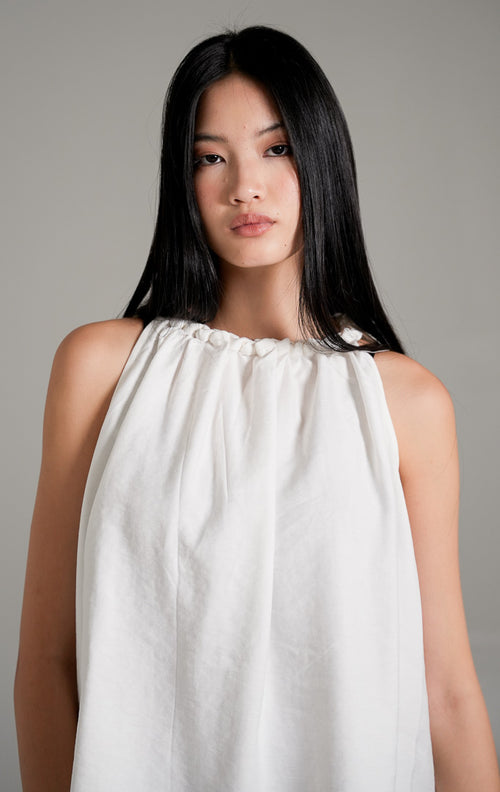 Keilani Top White, atasan tanpa lengan warna putih dengan kerah kerut kepang (braided neckline), dipadukan dengan celana jeans longgar biru.