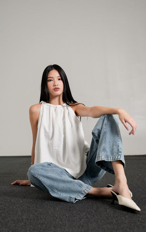 Keilani Top White, atasan tanpa lengan warna putih dengan kerah kerut kepang (braided neckline), dipadukan dengan celana jeans longgar biru.