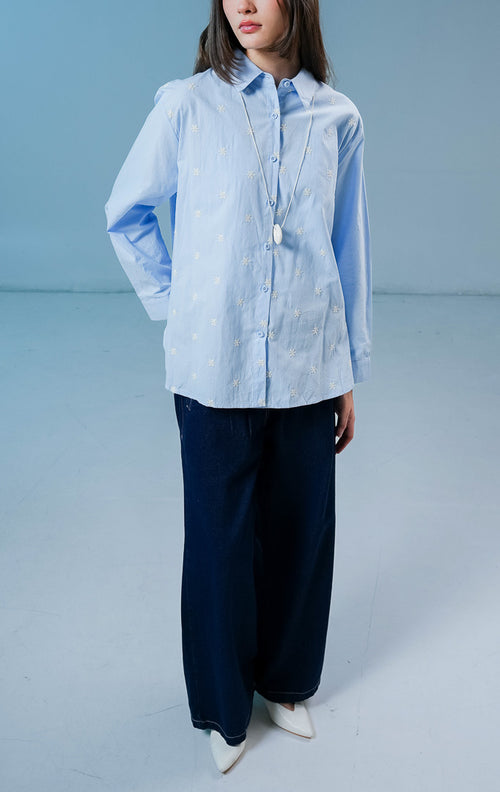 Model wanita mengenakan kemeja lengan panjang Ashford Top warna biru muda berbahan katun dengan detail kancing depan dan bordir bunga putih kecil.