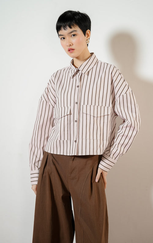 Model wanita mengenakan Georgette Classic Striped Collared Crop Shirt warna light pink, kemeja crop salur wanita lengan panjang berwarna merah muda dengan dua saku depan.