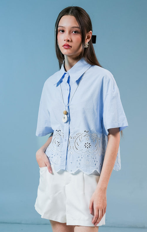 Model wanita mengenakan kemeja lengan pendek Annaleigh Top warna biru muda berbahan katun dengan detail bordir eyelet putih dan ujung bawah berpotongan scallop.