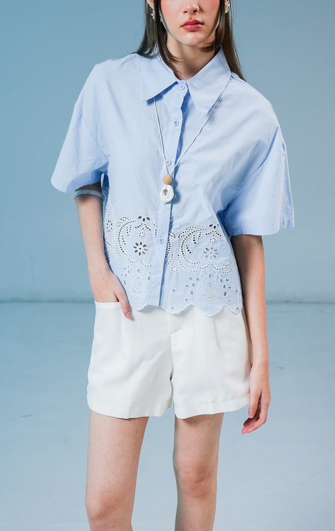 Model wanita mengenakan kemeja lengan pendek Annaleigh Top warna biru muda berbahan katun dengan detail bordir eyelet putih dan ujung bawah berpotongan scallop.
