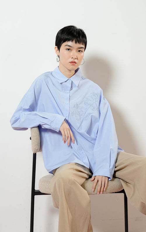 Detail dekat model mengenakan Zinnia Floral Embroidery Detail Shirt warna blue, atasan kemeja katun wanita lengan panjang warna biru muda dengan bordir motif bunga peony minimalis di dada.