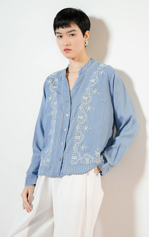 Model wanita mengenakan Brella Embroidery V-Neck Top warna blue denim, kemeja katun wanita lengan panjang berwarna biru muda dengan detail sulaman bordir bunga putih yang anggun. 