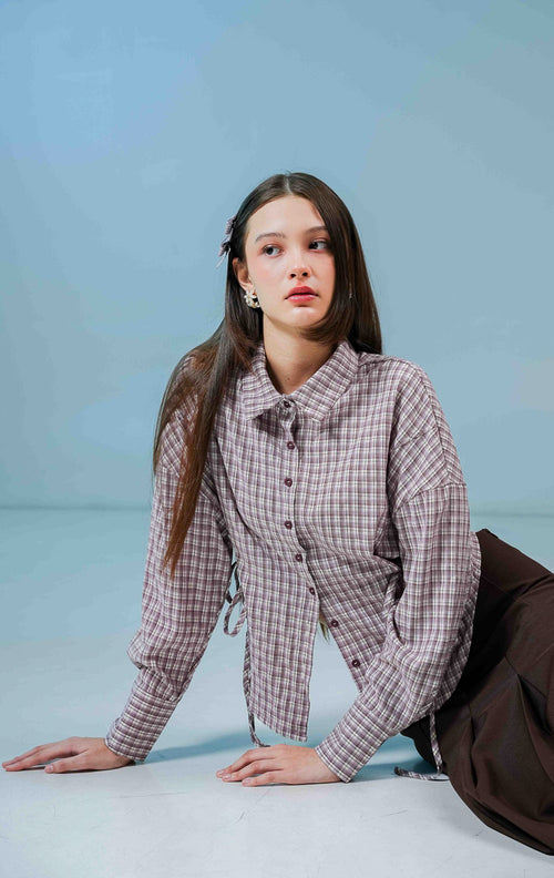Model wanita mengenakan Fawn Plaid Top warna ungu (purple), kemeja katun lengan panjang motif kotak-kotak klasik dengan detail tali serut di bagian samping. 
