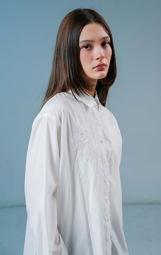 Model wanita mengenakan kemeja lengan panjang Cecilina Top warna putih bersih berbahan katun dengan detail kancing depan dan bordir bunga yang elegan di bagian dada. 