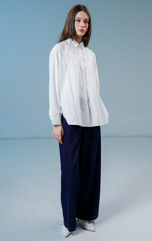Model wanita mengenakan kemeja lengan panjang Cecilina Top warna putih bersih berbahan katun dengan detail kancing depan dan bordir bunga yang elegan di bagian dada. 