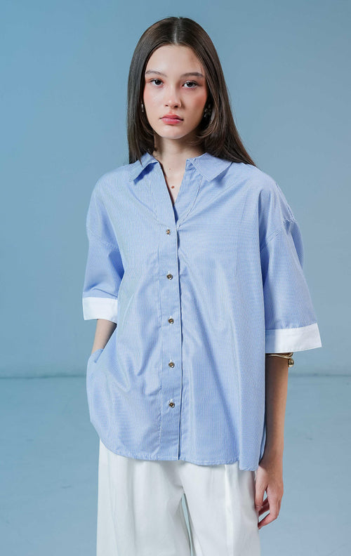 Model wanita mengenakan Naudre Stripe Contrast Hem Top warna blue, kemeja salur wanita motif garis biru-putih dengan detail ujung lengan putih kontras yang modern. 