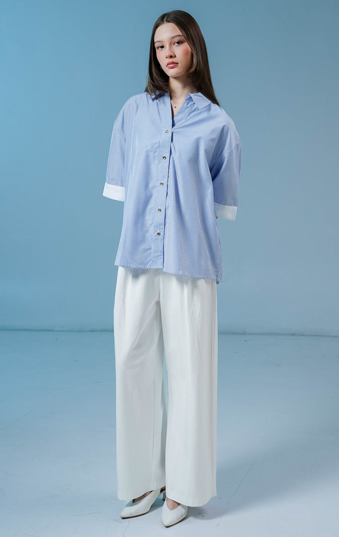 Model wanita mengenakan Naudre Stripe Contrast Hem Top warna blue, kemeja salur wanita motif garis biru-putih dengan detail ujung lengan putih kontras yang modern. 
