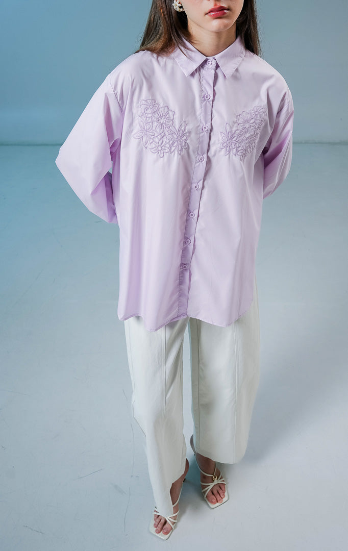 Model wanita mengenakan kemeja lengan panjang Cecilina Top warna ungu muda atau lilac berbahan katun dengan detail kancing depan dan bordir bunga yang elegan di bagian dada. 
