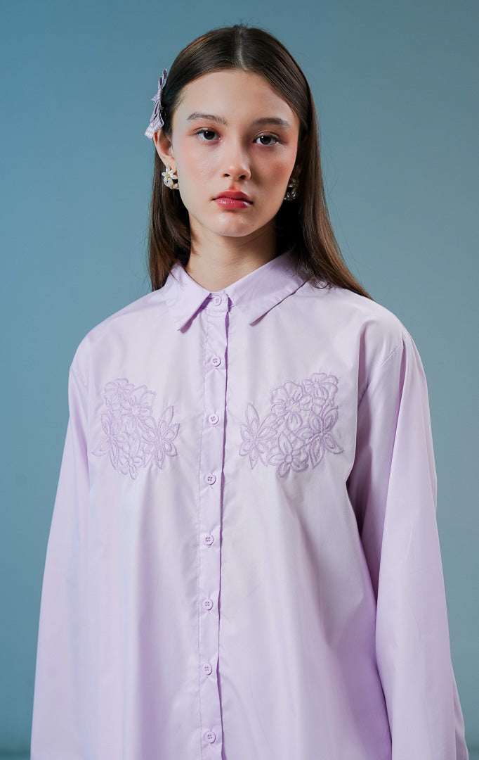 Model wanita mengenakan kemeja lengan panjang Cecilina Top warna ungu muda atau lilac berbahan katun dengan detail kancing depan dan bordir bunga yang elegan di bagian dada. 