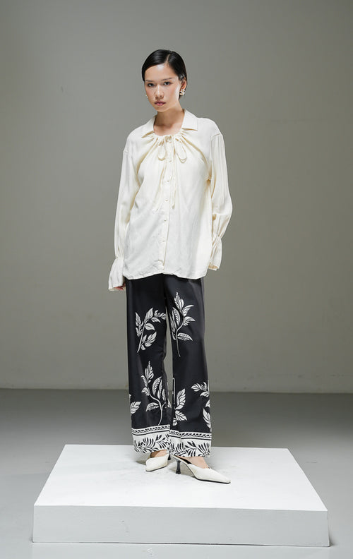 Blouse wanita Kensley Top warna ivory krem muda lengan panjang dengan detail tali pita leher (tie neck), kerah kemeja, dan kancing depan. 