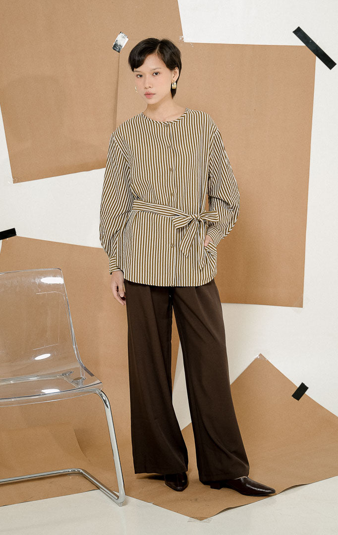 Model berdiri mengenakan Kiano Waist Tie Stripe Shirt Brown, kemeja salur wanita lengan panjang motif garis cokelat-putih dengan detail tali ikat di pinggang dipadukan celana kulot panjang.