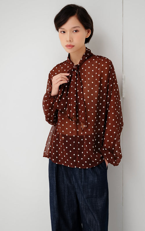 Atasan chiffon wanita transparan (sheer) warna cokelat tua (dark brown) Lillith Top motif polkadot dengan aksen ikatan leher (tie-neck) dan lengan panjang. 
