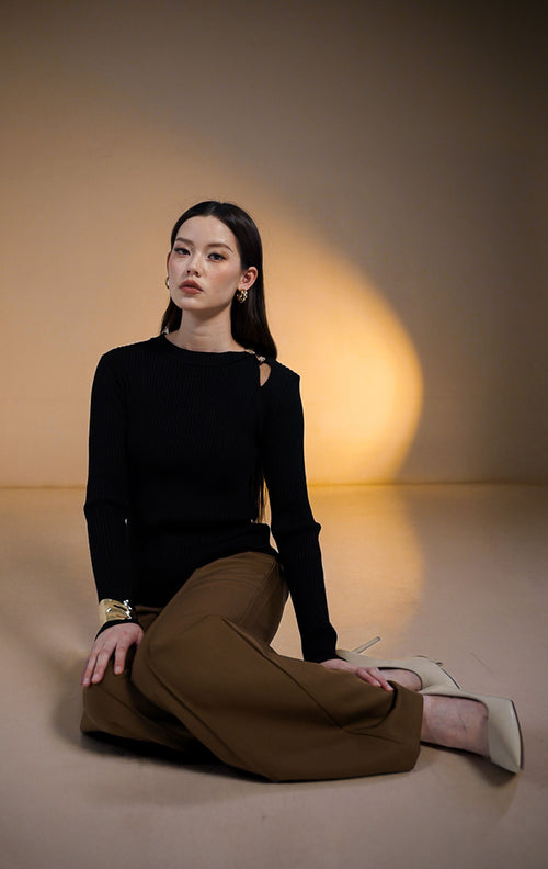 Model wanita mengenakan Loraine Ribbed Knit Top warna hitam, atasan rajut lengan panjang tekstur alur dengan detail lubang (cut-out) di bahu kiri yang dihiasi bros emas, dipadukan dengan celana cokelat.