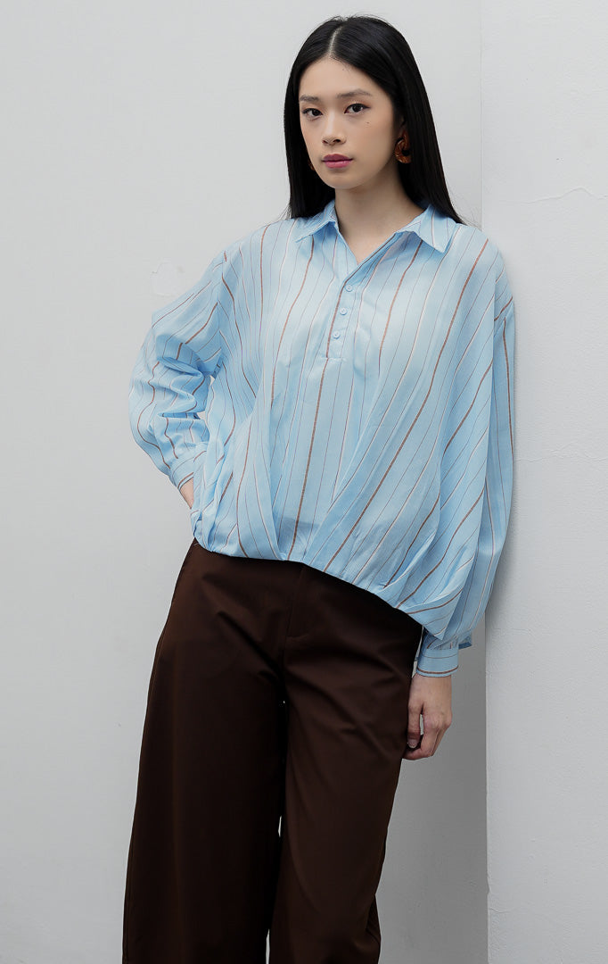 Kemeja wanita lengan panjang warna biru muda (light blue) Lorena Top dengan aksen siluet lipat silang (hi-low crossover hem), bahan katun ringan bermotif garis salur, dan bukaan kancing depan.