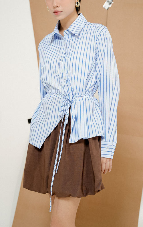 Model berdiri mengenakan Louella Tie-Front Stripe Shirt Light Blue, kemeja salur wanita lengan panjang warna biru muda motif garis vertikal dengan aksen tali ikat di pinggang. 