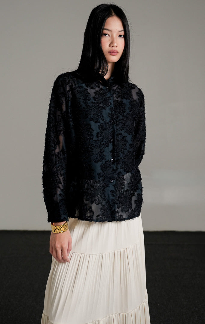 Loui Top Navy, kemeja lengan panjang warna biru tua semi-transparan dengan detail tekstur bulu halus, dipadukan dengan rok panjang lipit putih gading.
