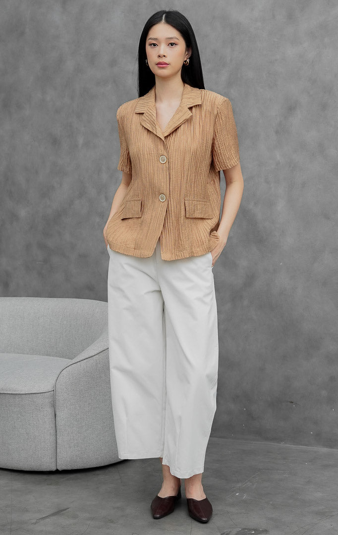 Blazer wanita lengan pendek warna cokelat muda (light brown) Lovegood Blazer berbahan crinkle polyester dengan tekstur plissé, kerah notch lapel, bukaan kancing depan, dan saku saku penutup (flap pockets).